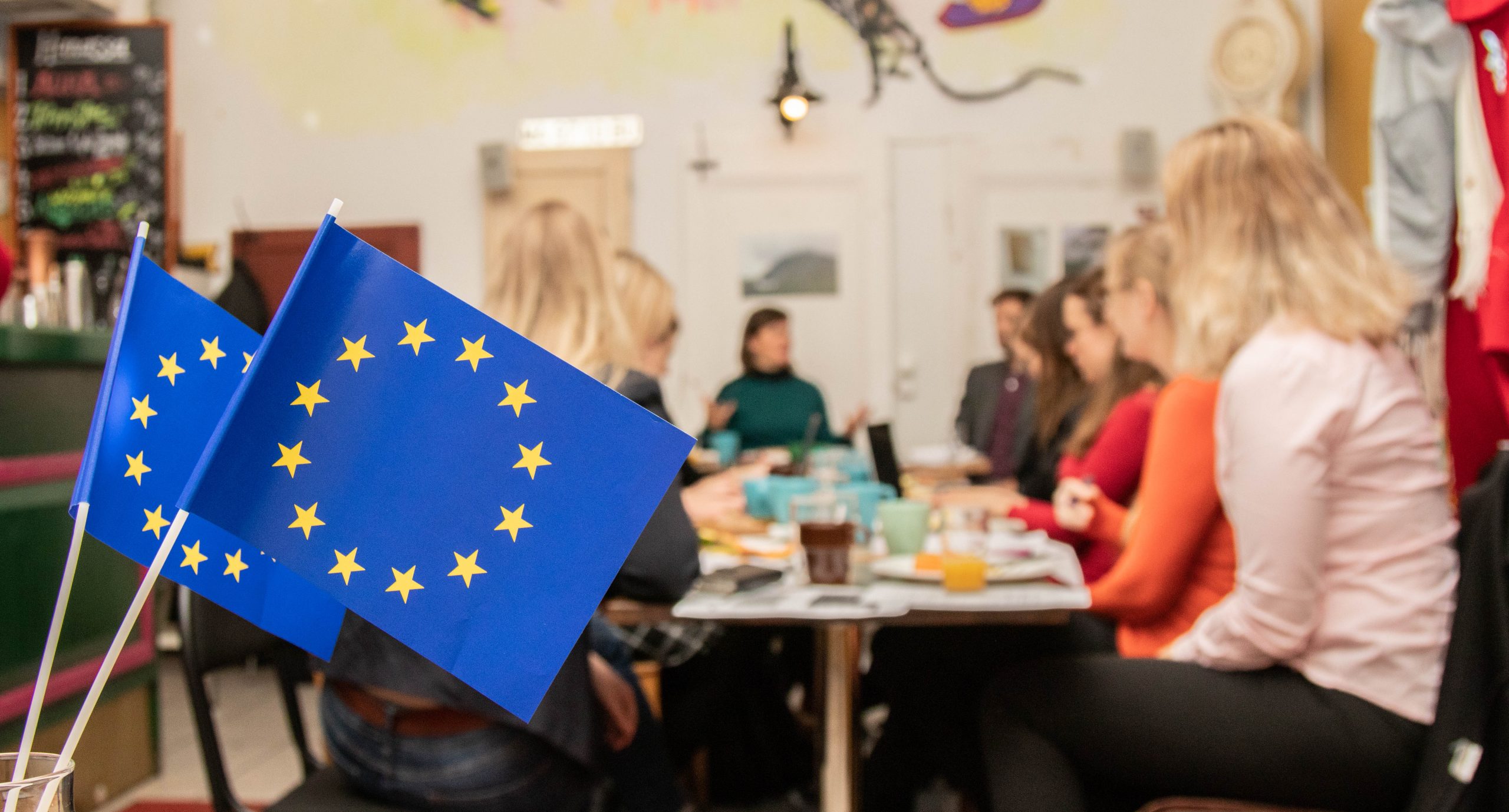 Kuvassa on kaksi pientä EU-lippua ja taustalla ihmisiä pöydän ympärillä.