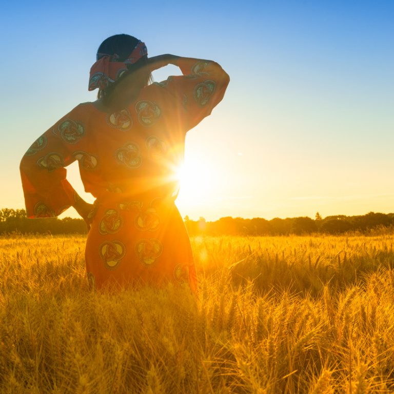 Nainen seisoo viljapellolla ja katsoo auringonnousuun.