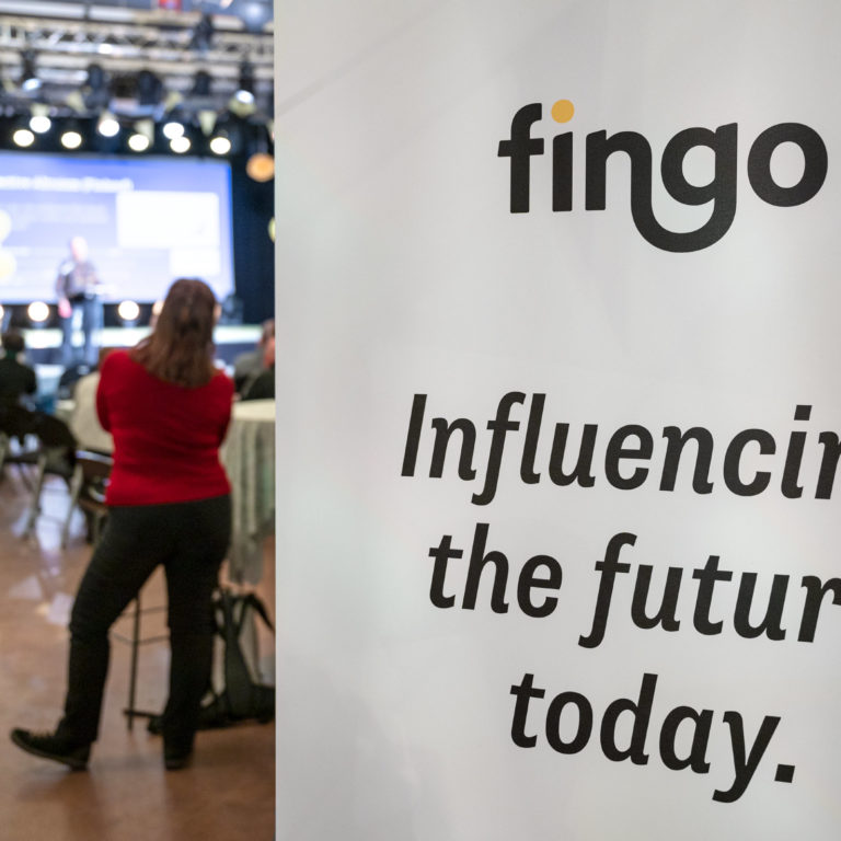 Fingo roll-up, jossa Fingon logo ja teksti 'Influencing the future today.' ja taustalla kokoustilan lava ja osallistujia.