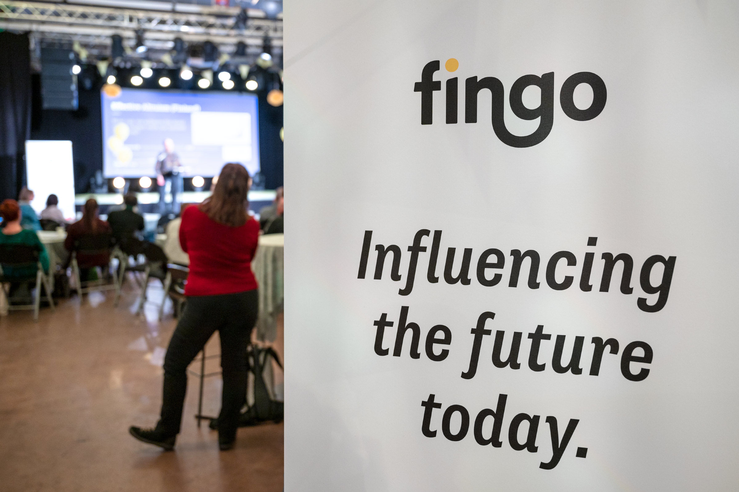 Fingo roll-up, jossa Fingon logo ja teksti 'Influencing the future today.' ja taustalla kokoustilan lava ja osallistujia.