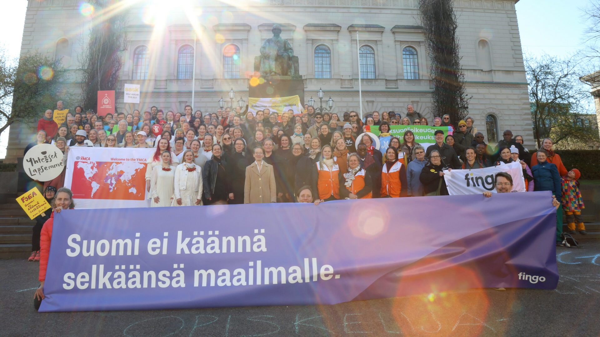 Iso ihmisjoukko poseeraa kampanjabannereiden kanssa Suomen Pankin edustalla.