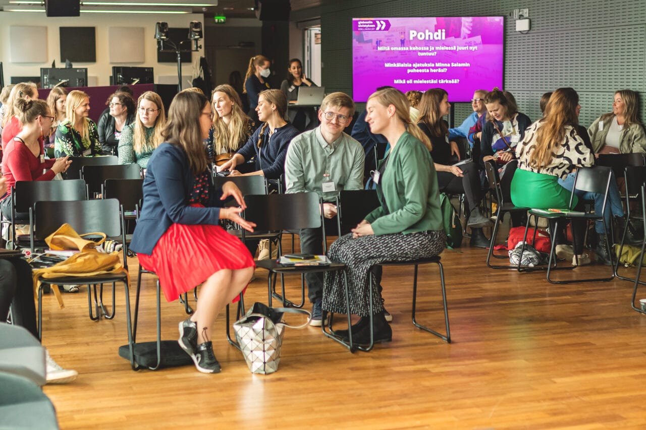 Seminaarin yleisö istuu tuoleilla ja keskustelee keskenään.|Kuohuviinilaseja pöydällä.|Paneelikeskustelijat istuvat tuoleilla.||Fingon roll-up