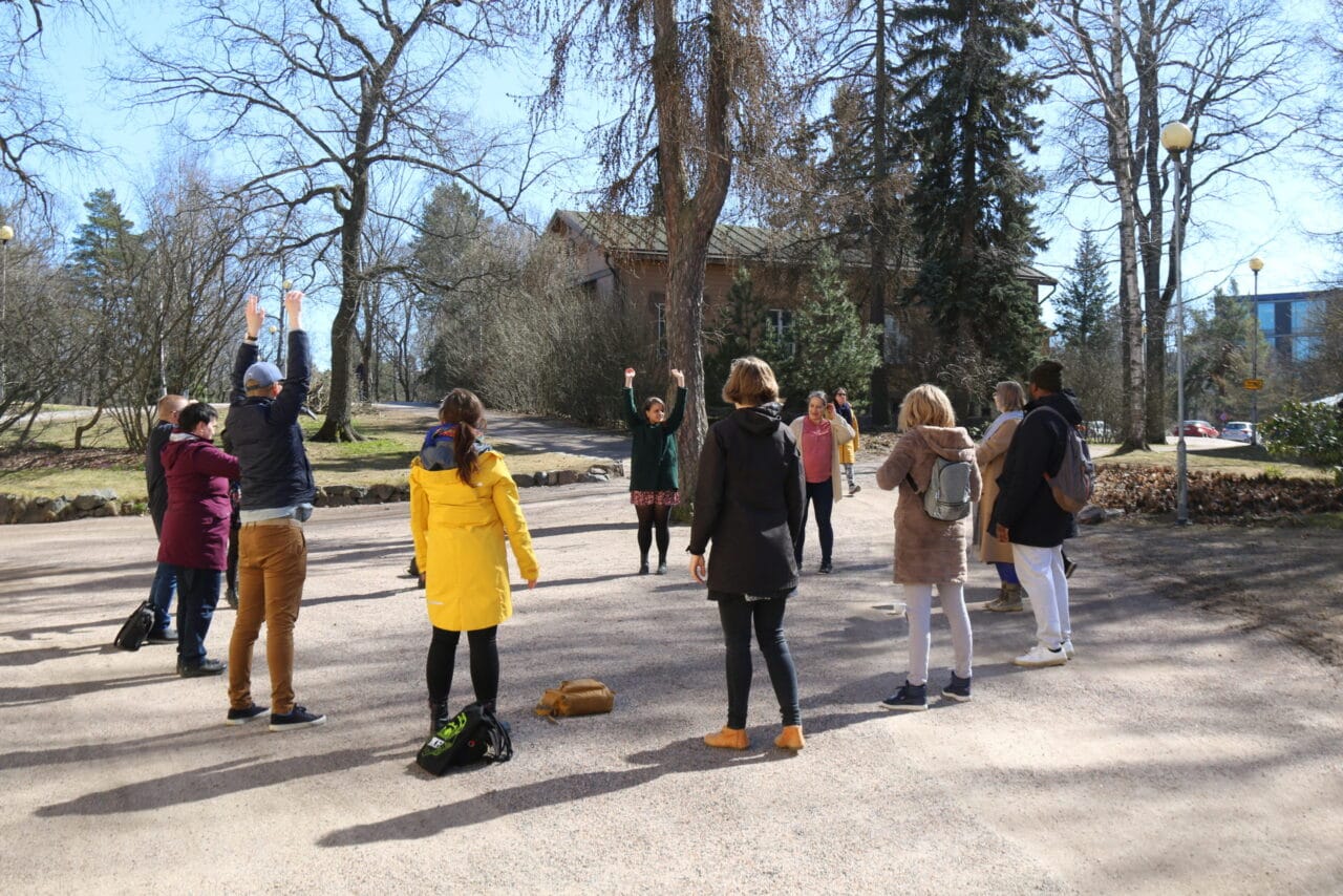 Hackathon osallistujat seisovat ringissä hiekkakentällä. Aurinko paistaa.