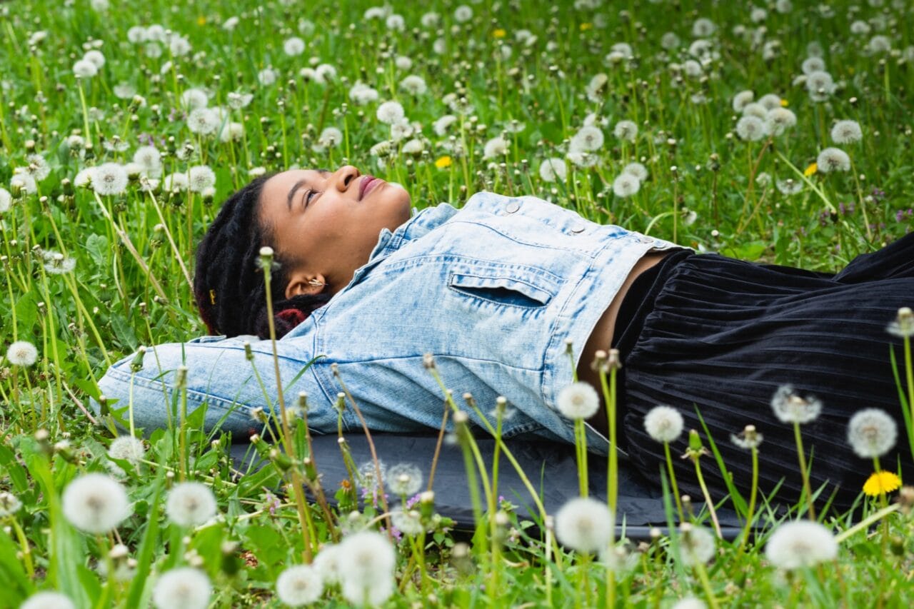 Henkilö makoilee voikukkaniityn keskellä ja katselee taivaalle.