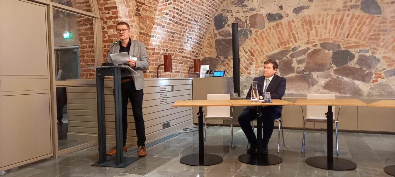 |Viisi henkilöä istuu pöytien äärellä puoliringissä.