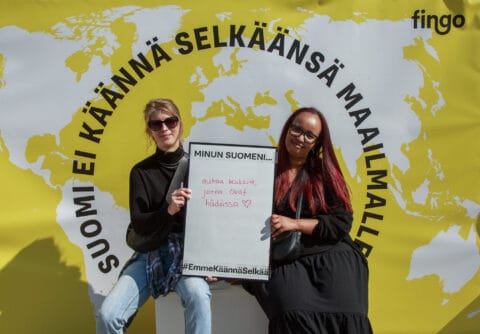 Kaksi henkilöä pitää välissään taulua, jossa teksti: Minun suomene auttaa kaikkia jotka ovat hädässä. Taustalla maailmankartta ja Fingon logo.